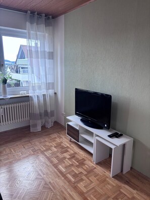 A room with a television on a white stand, a radiator, and a window with sheer curtains.