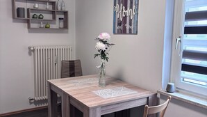 Een eethoek met een houten tafel, stoelen, een vaas met bloemen, een radiator, een plankje met decoratieve objecten, en een raam met jaloezieën.