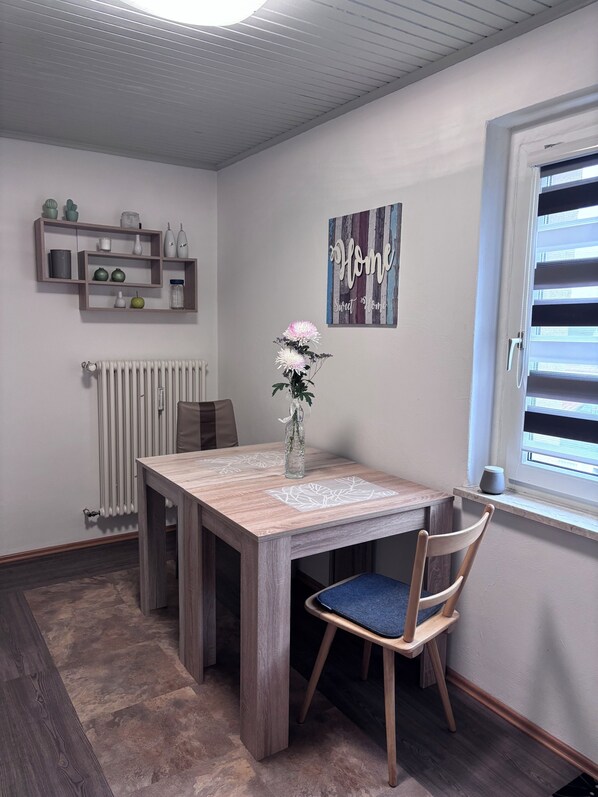 A dining area with a wooden table, chairs, a vase with flowers, a radiator, a shelf with decorative items, and a window with blinds.