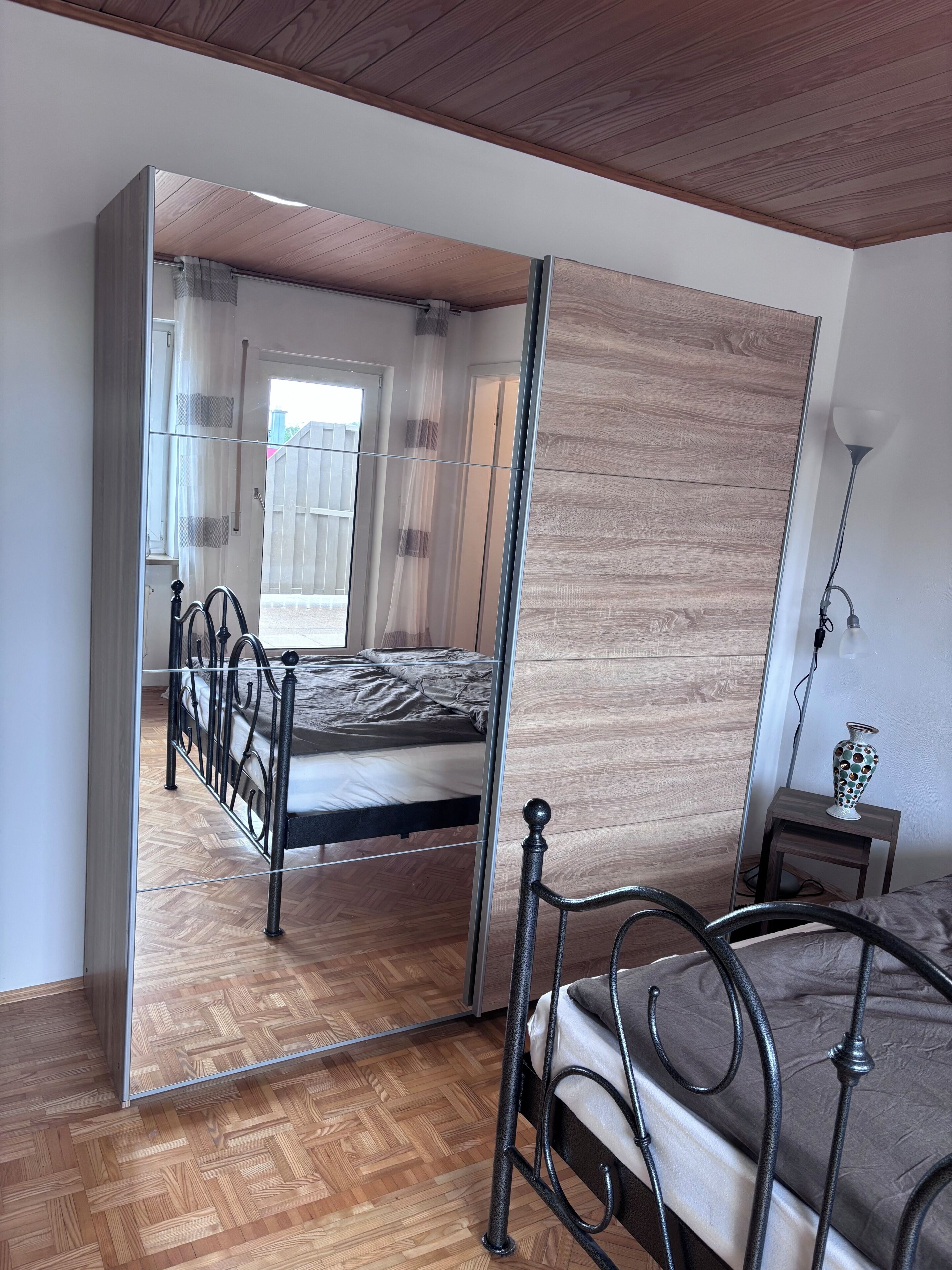 A bedroom with a bed, a mirror, a small table with a lamp, and a wooden floor.