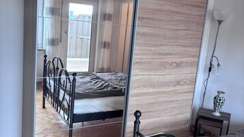 A bedroom with a bed, a mirror, a small table with a lamp, and a wooden floor.
