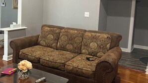A living room with a brown patterned sofa, a glass coffee table with a vase and remote controls, a ceiling fan, and a white wall with a picture frame. - World Cup Gem 12 minutes from all attractions (Kansas City)