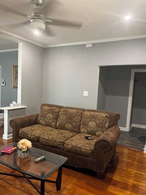 Ein Wohnzimmer mit einer braunen, gemusterten Couch, einem Glastisch mit einer Vase und Fernbedienungen, einem Deckenventilator und einer weißen Wand mit einem Bilderrahmen.