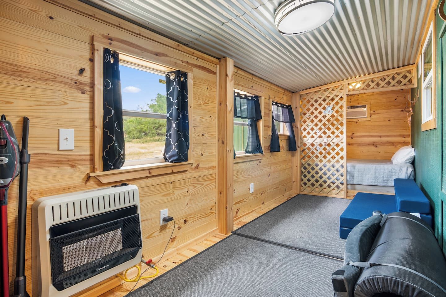 A compact RV interior with two windows, a heater, a bed, and a small seating area.