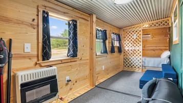 A compact RV interior with two windows, a heater, a bed, and a small seating area.