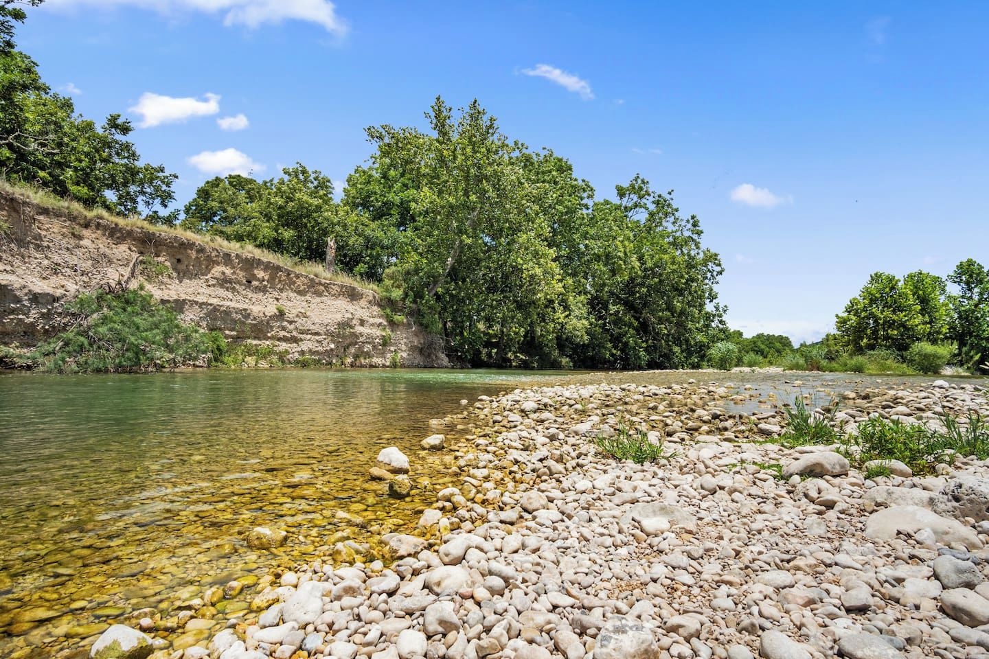 A riverbank with clear water, pebbles, and dense green trees.
