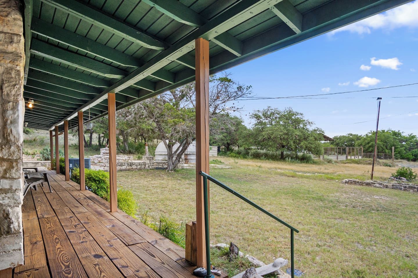A covered outdoor area with a wooden deck, stone walls, and a view of a grassy landscape with trees.