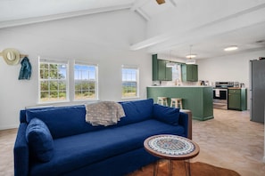 A living room with a blue sofa, a small round table, and a kitchen area with green cabinets and a black refrigerator.