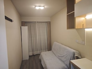 Een kleine, smalle kamer met een bed, een witte kledingkast, een spiegel en een lamp aan het plafond.
