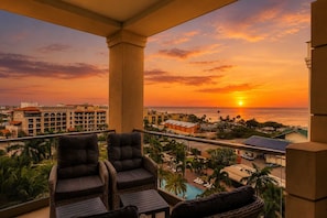 Ein Balkon mit Korbstühlen und Pool mit Blick auf eine Küstenlandschaft, einschließlich Gebäude und Palmen, im Sonnenuntergang.