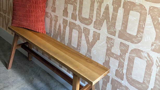 Un banco de madera con un cojín rojo frente a una pared con patrones de texto repetidos.