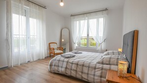 Ein Schlafzimmer mit einem Bett, einem Sessel, einem Nachttisch mit Lampe und einem Fenster mit Vorhängen.