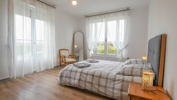A bedroom with a bed, a chair, a bedside table with a lamp, and a window with curtains.