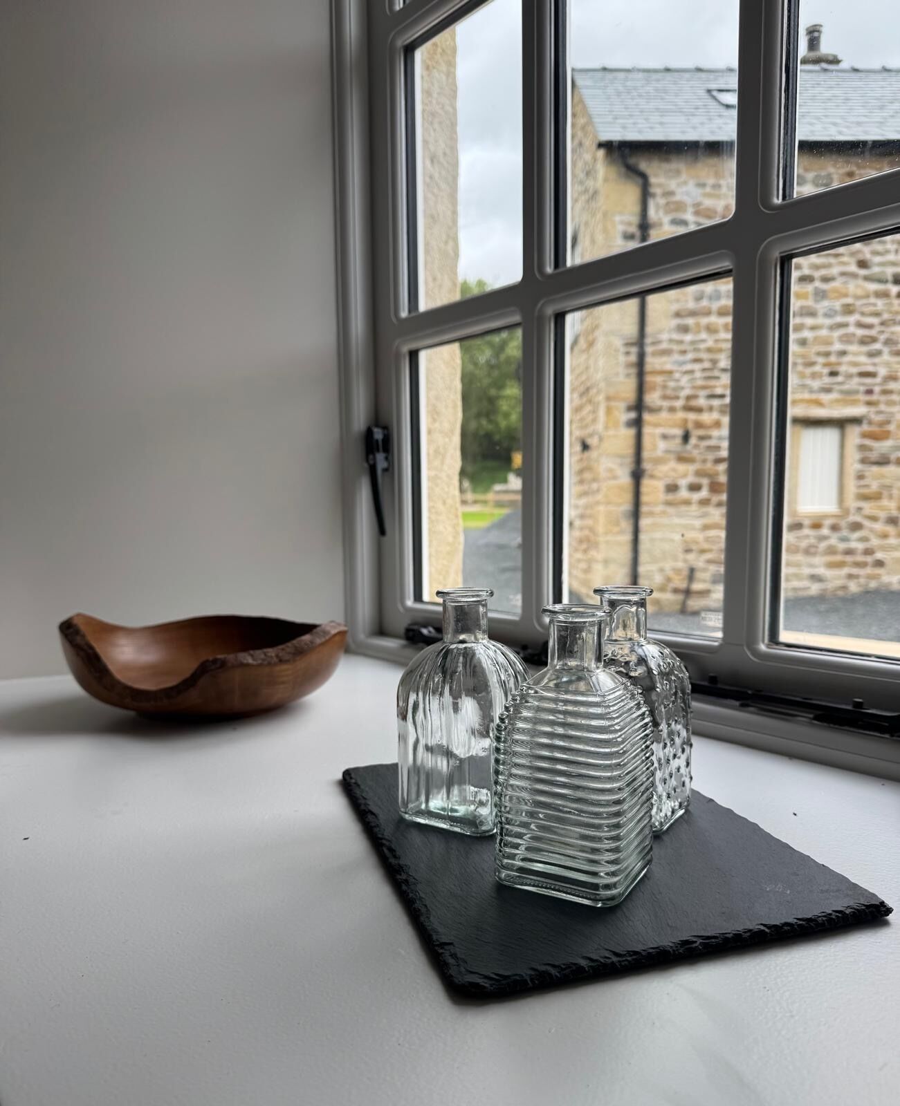 A window with a view of stone buildings, a wooden bowl, and three glass bottles on a black slate.
