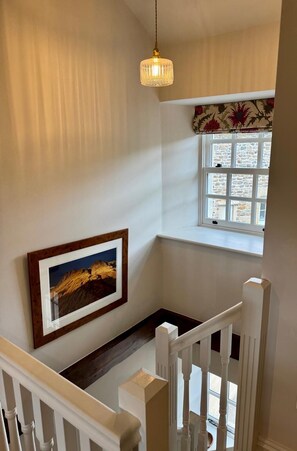 Un escalier avec des rampes blanches, un tableau encadré au mur, un luminaire suspendu et une fenêtre agrémentée d’une lambrequin à motifs floraux.