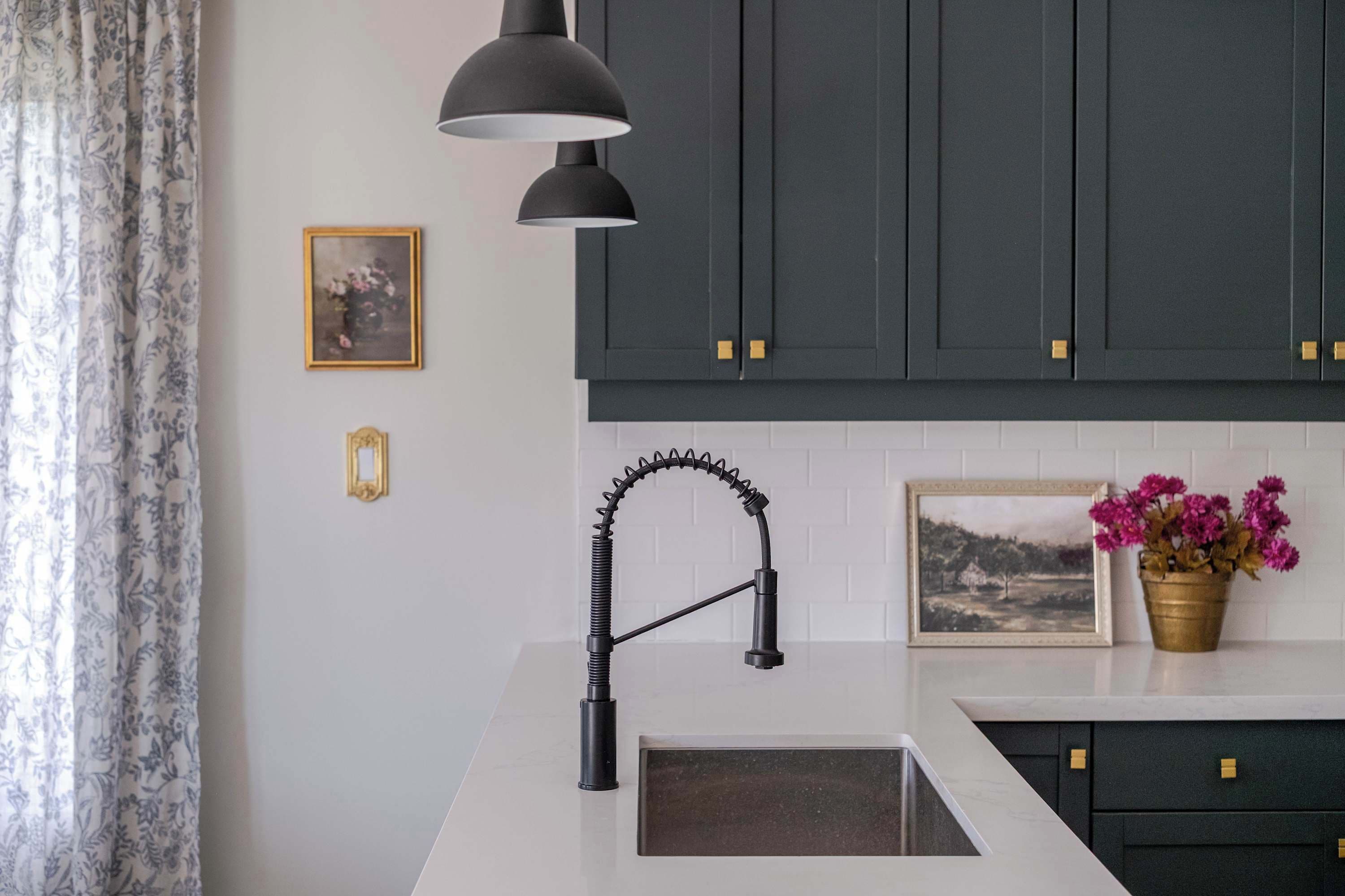 Une cuisine avec des meubles de couleur sombre, un plan de travail blanc, un robinet noir et un luminaire suspendu.