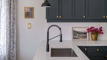 Une cuisine avec des meubles de couleur sombre, un plan de travail blanc, un robinet noir et un luminaire suspendu.