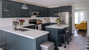 Una cocina moderna con gabinetes oscuros, encimeras blancas, electrodomésticos de acero inoxidable e iluminación colgante.