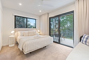 Een slaapkamer met een groot bed, een nachtkastje met een lamp, een raam en een schuifdeur die leidt naar een buitenruimte met stoelen.