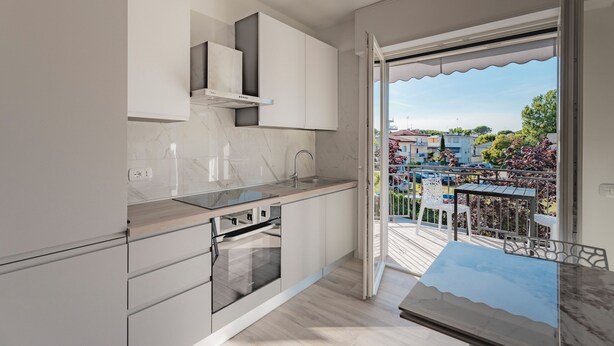 A modern kitchen with a built-in oven, a sink, and a window overlooking a balcony with outdoor furniture. - Sundown Living | New & Modern Apartment | 200 m from the beach | Balcony | Air conditioning (Lignano Sabbiadoro)