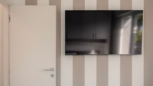 A modern room with a flat-screen TV mounted on a wall with vertical stripes, a white door, and wooden flooring. - Sundown Living | New & Modern Apartment | 200 m from the beach | Balcony | Air conditioning (Lignano Sabbiadoro)