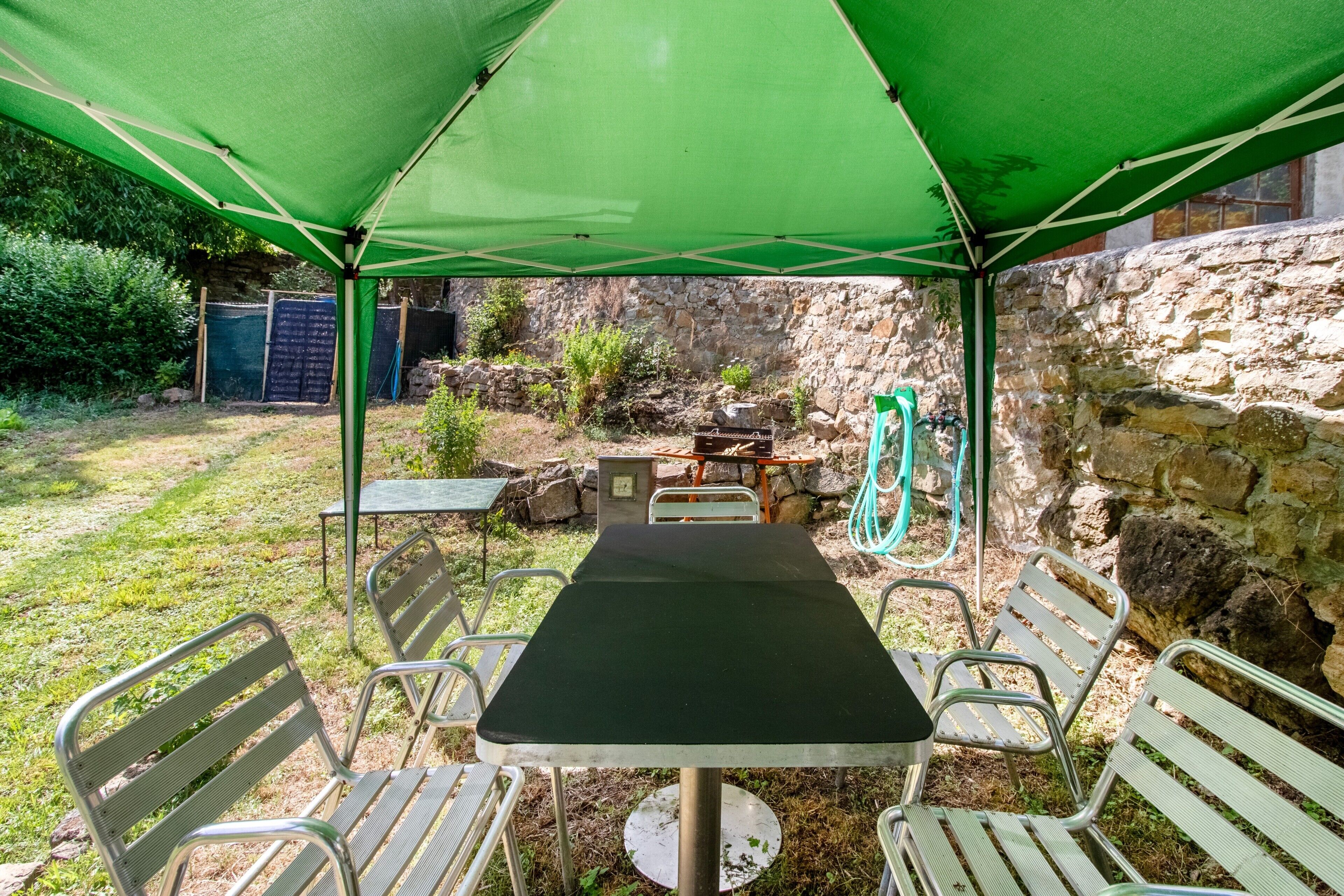 A covered outdoor seating area with a table and chairs, a stone wall, and a hose in the background.