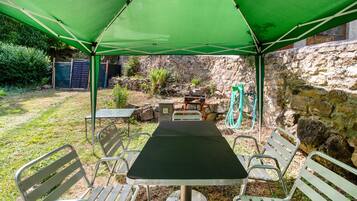 A covered outdoor seating area with a table and chairs, a stone wall, and a hose in the background.