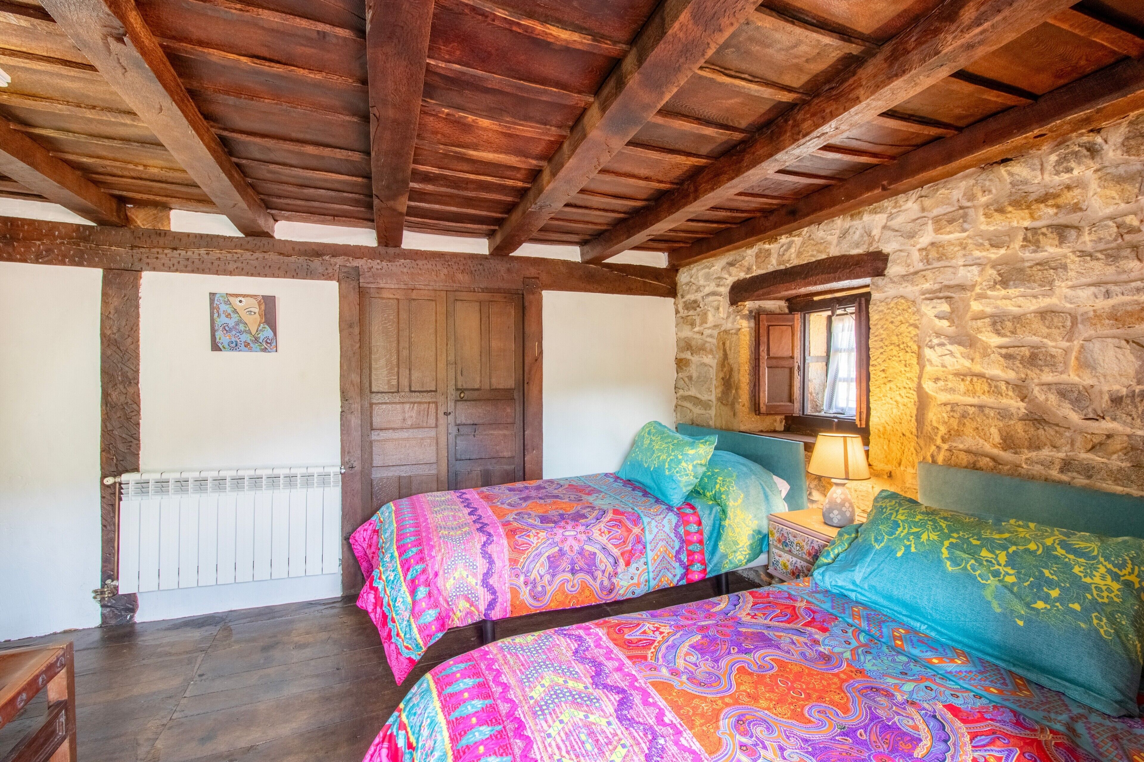 Ein Zimmer mit einer Steinwand, Holzbalken an der Decke, einem Bett mit bunter Bettwäsche, einer Holztür und einem Fenster mit Aussicht.