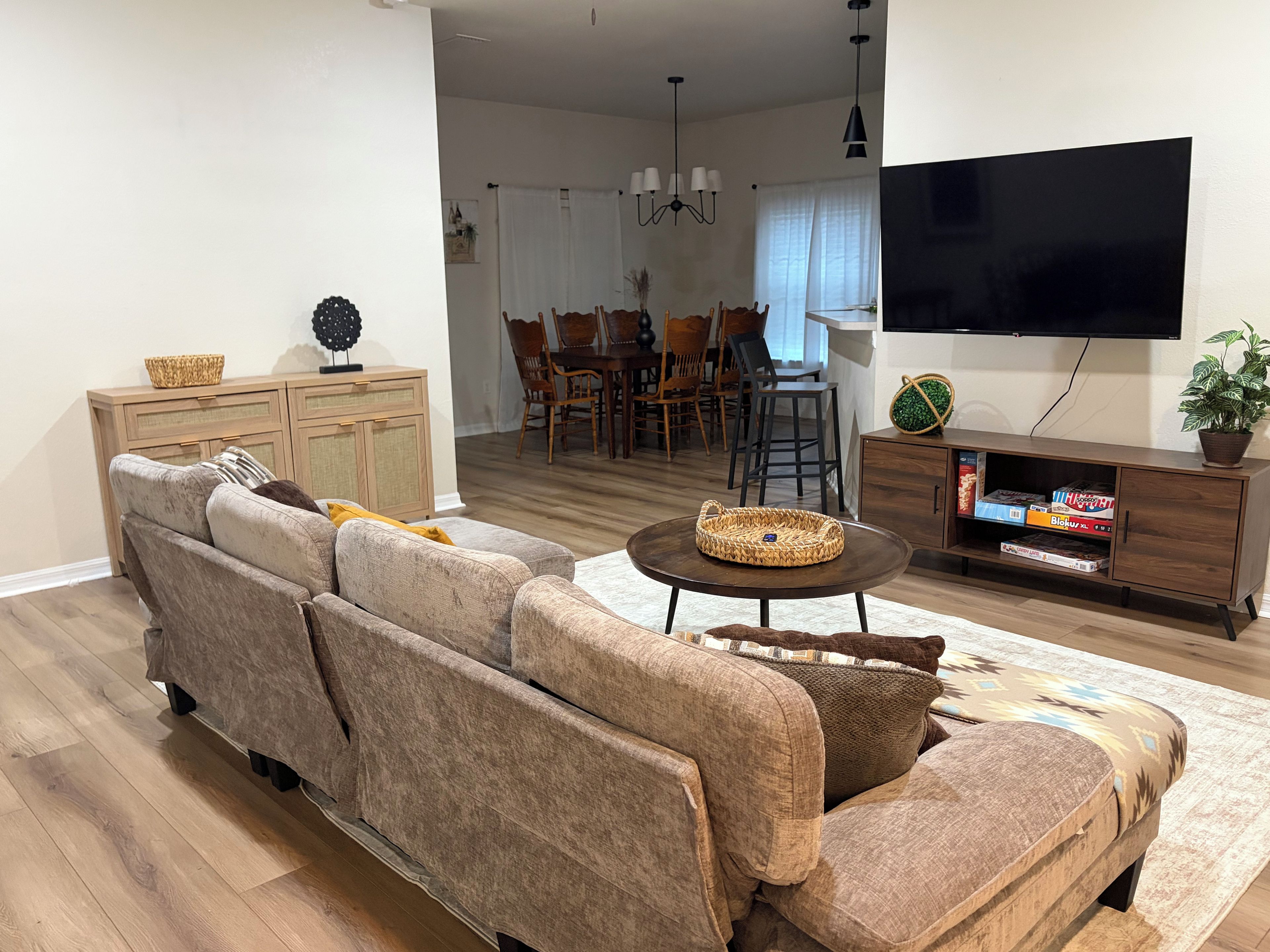 Uma sala de estar com sofá seccional, mesa de centro, televisão de tela plana e área de jantar com cadeiras e mesa.