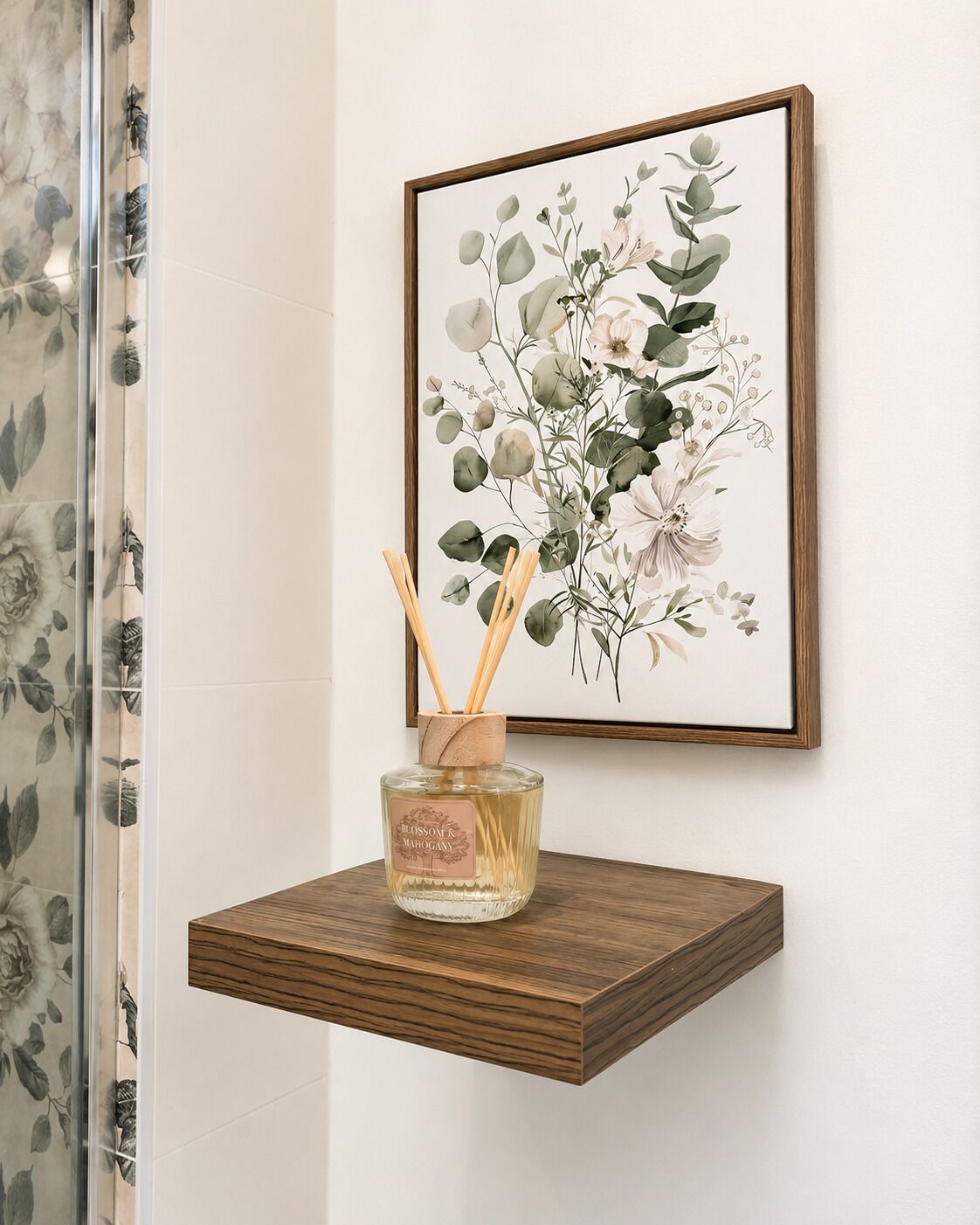 Een ingelijst botanisch kunstwerk aan een muur, een houten plank met een parfumfles, en een douchegordijn van glas met een bloemendesign.