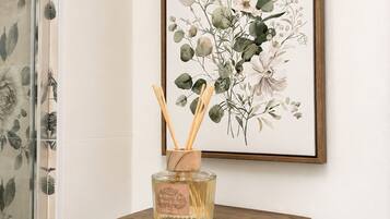 Een ingelijst botanisch kunstwerk aan een muur, een houten plank met een parfumfles, en een douchegordijn van glas met een bloemendesign.