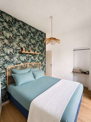 A bedroom with a bed, a wooden headboard, a wall with a leafy pattern, a hanging light fixture, and a shelf with towels. - Cozy apartment in the heart of Saumur – Château & Loire on Foot (Saumur)