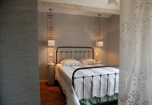 Un dormitorio con estructura de cama metálica, una cama individual con cubrecama a rayas, dos lámparas de noche y una pared de ladrillo.