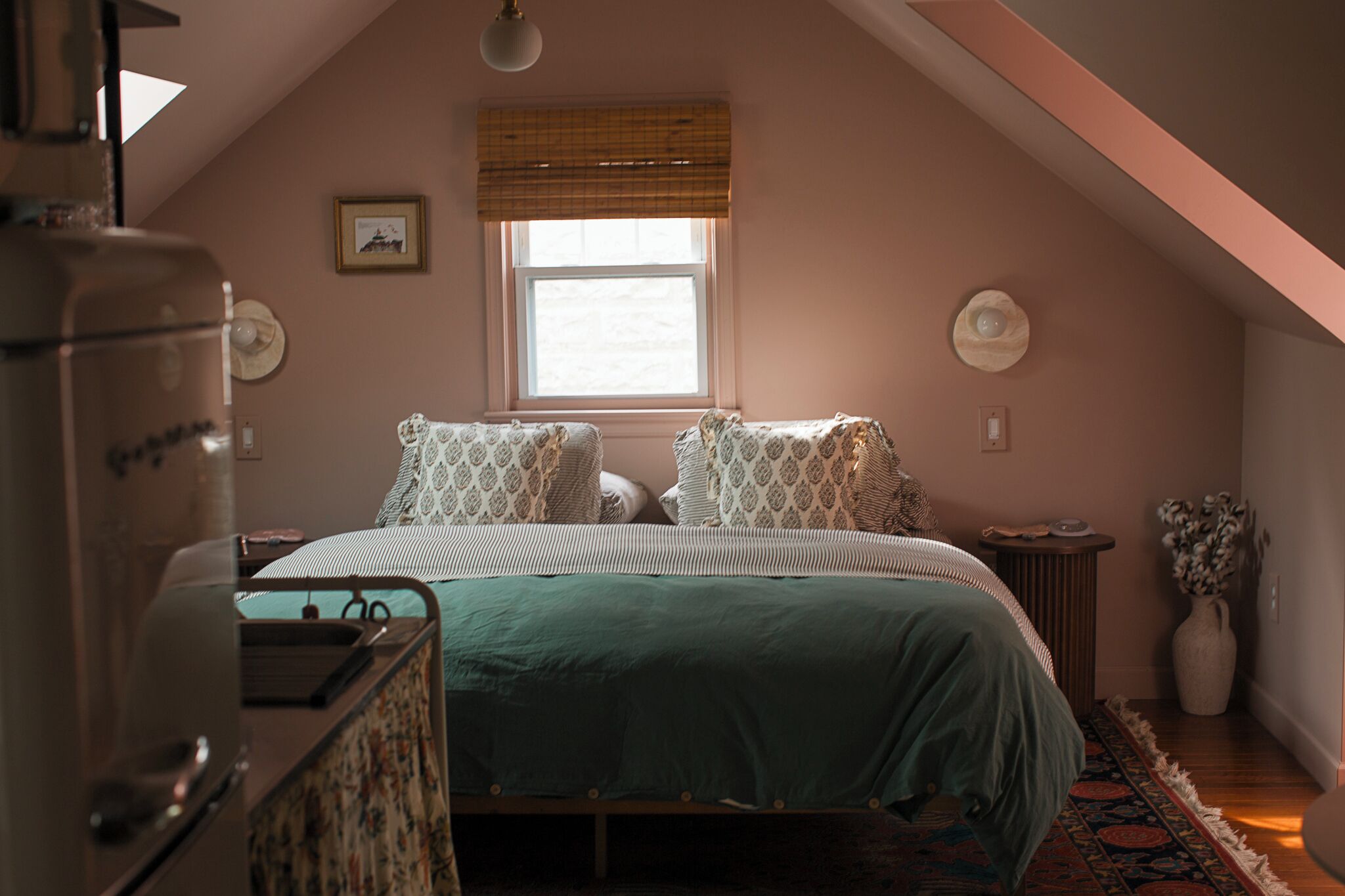 Un dormitorio con cama, cómoda, ventana con persianas, un marco de foto y un jarrón con flores.