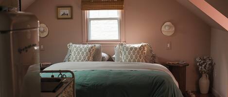 Un dormitorio con cama, cómoda, ventana con persianas, un marco de foto y un jarrón con flores.