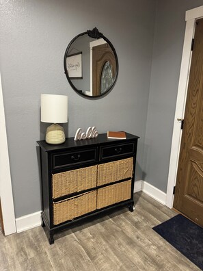 A black console with wicker baskets, a white lamp, and a mirror with a 'Hello' sign on a gray wall.