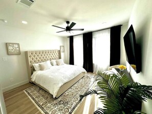 A bedroom with a tufted headboard, a bed with a patterned bedspread, a ceiling fan, a large window with curtains, a flat-screen TV mounted on the wall, and a potted plant. - Prime UTSW | 3BR Home + Patio+Garage (Dallas)