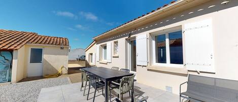 A patio area with a table and chairs, a white building with windows, and a tiled roof.