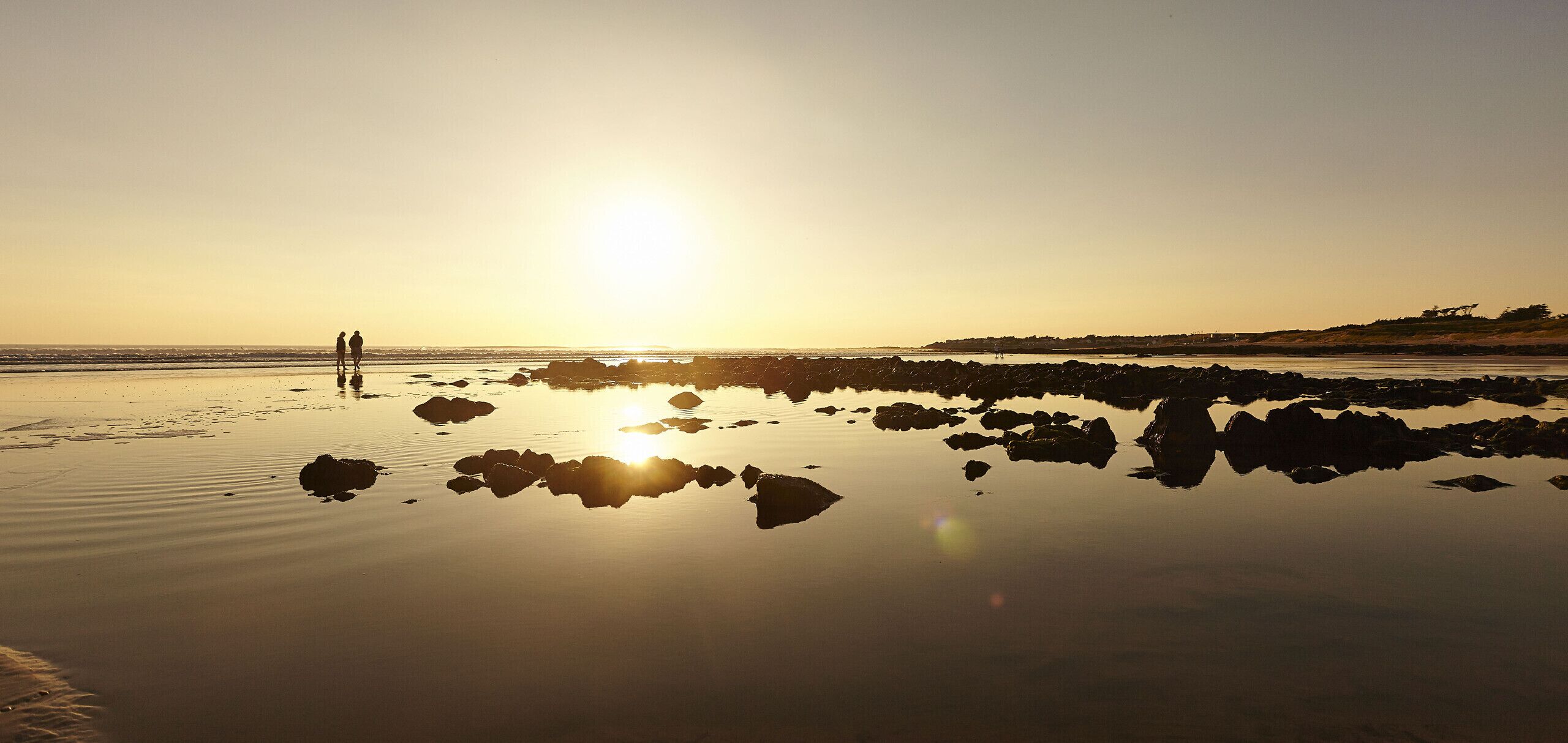 A beach scene with calm water, rocks, and two people walking.
