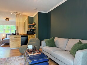 A modern living room with a sofa, a dining area with a microwave, and a kitchen with wooden shelves.