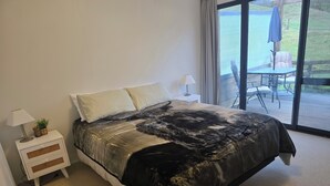Et soveværelse med en stor seng, et natbord med lampe, og udsigt til en udendørs terrasse.