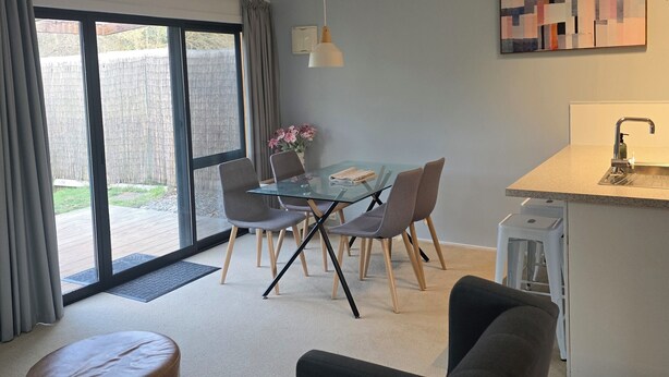 En moderne stue med et glasbord, grå stole, en sofa og et køkkenområde med vask og bordplade.