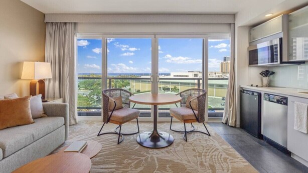 A hotel room with a balcony, a sofa, a dining table with chairs, a kitchen area, and a large window with a city view. - Ala Moana 1 Bedroom · Ocean Views, Beach, Mall (Honolulu)