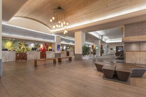 A spacious lobby with a wooden floor, a reception desk, seating areas, and a modern chandelier.