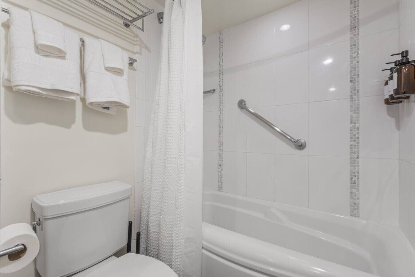 Un baño con inodoro, ducha con barra de apoyo y toallero con varias toallas.