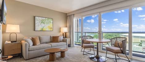 Habitación de hotel con sofá, dos mesitas auxiliares, una mesa de centro, una silla y un balcón con vistas.