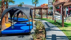 Area piscina con lettini blu, una zona relax coperta e un vialetto fiancheggiato da piante.