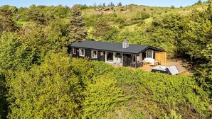 Une maison moderne avec un toit noir, entourée d’une végétation luxuriante et avec une colline en arrière-plan.