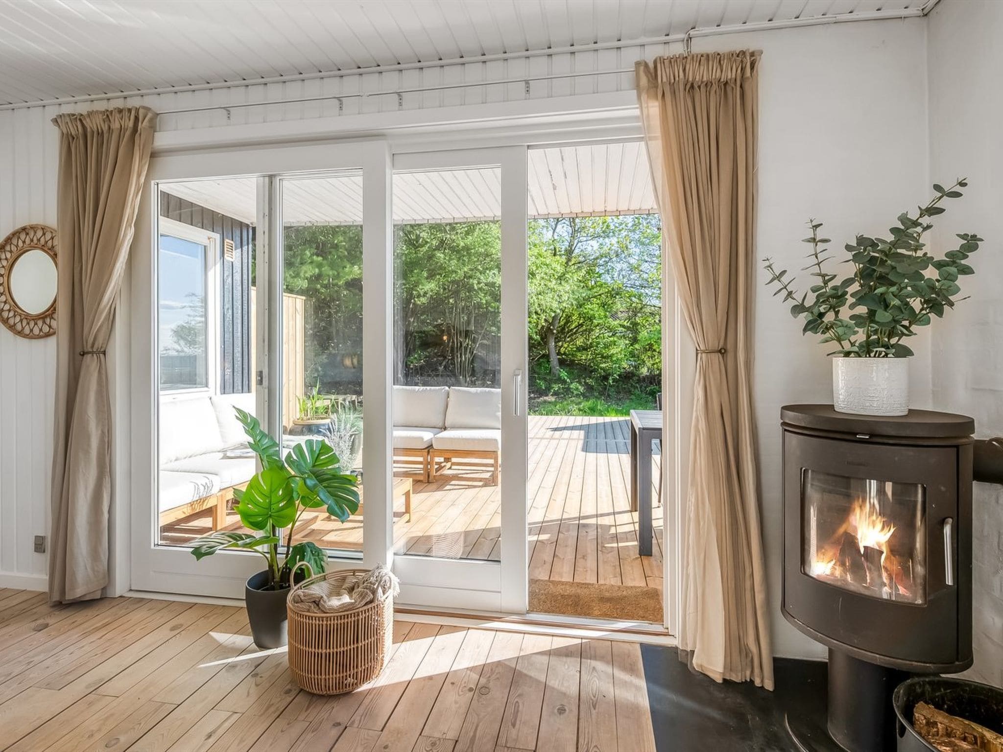 A room with a wooden floor, a sofa, a large window with curtains, a fireplace, and a view of an outdoor deck.