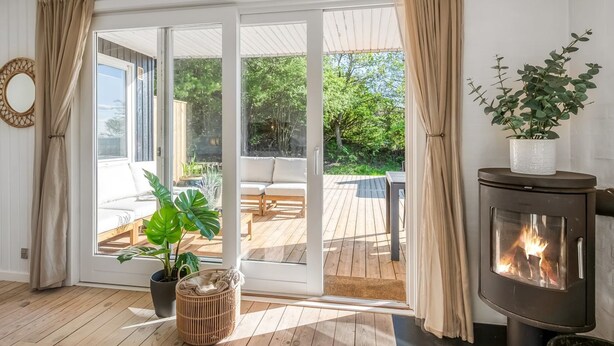 Une pièce dotée d’un plancher en bois, d’un canapé, d’une grande fenêtre avec des rideaux, d’une cheminée et donnant sur une terrasse extérieure.